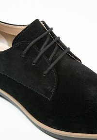 Chaussure habillée en daim noir présentant une texture lisse, un bout arrondi et un design à lacets. Semelle en caoutchouc avec un subtil accent tan le long du bord.