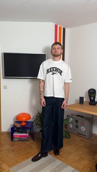 Valge lühikeste varrukatega T-särk musta "REEBOK" logoga, kombineeritud lahtiste tumedate teksapükste ja tumedate jalanõudega. Lihtne, kaasaegne disain.