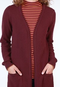 Burgundy knitted cardigan with a textured diamond pattern, open front, and patch pockets, layered over a striped turtleneck in warm colors.