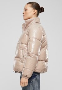 Puffy, ribbet jakke i lys beige med glansfull finish, høy krage og sidelommer; har elastiske mansjetter og cropped design.