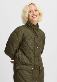 Manteau matelassé vert olive avec un col haut, des boutons pression et des poches. Présente un motif de couture en diagonale et une texture lisse.