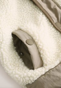 Gros plan sur une poche beige à bouton-pression sur un tissu polaire blanc texturé, montrant une surface douce semblable à de la laine et une doublure lisse à l'intérieur de la poche.