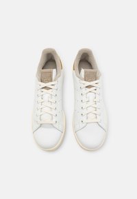 Biele kožené tenisky s textúrovanou béžovou podšívkou, plochými šnúrkami a jemnými perforáciami. Obsahujú malý zlatý akcent na päte.
