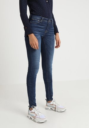 Persona vistiendo jeans ajustados azul oscuro, zapatillas blancas con rosa claro y una camisa de manga larga azul oscuro con pequeños puntos blancos.