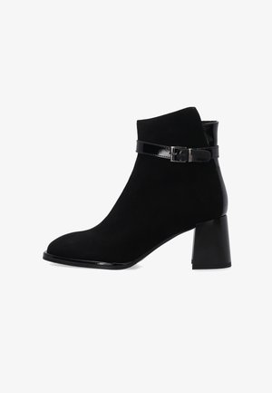 Schwarzer Knöchelstiefel aus Wildleder mit einem glänzenden schwarzen Schnallenriemen. Hat eine abgerundete Zehenpartie und einen stabilen Blockabsatz.