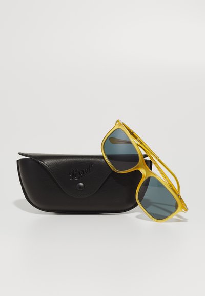 Óculos de sol amarelos com lentes cinzas ao lado de uma capa de pele preta, apresentando um logotipo em relevo e um fecho de pressão.