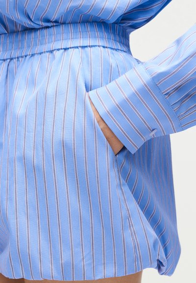 Κοντινό πλάνο στο χέρι ενός ατόμου στην τσέπη μπλε και λευκών ριγωτών σορτς με ελαστική μέση, σε συνδυασμό με ένα αντίστοιχο μακρυμάνικο πουκάμισο.