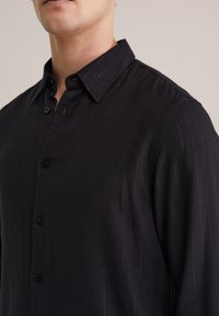 Mustmust valge triibuline särk, millel on struktuurne krae ja tekstureeritud kude siledal viimistlusel. Ees on neli nähtavat nööpi.