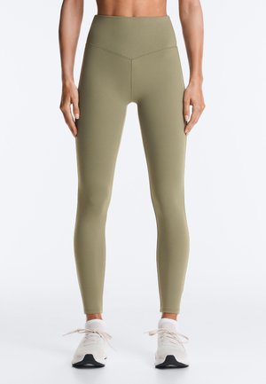 Person iført olivengrønne leggings med høj talje og hvide sneakers, stående med armene afslappet langs siden mod en ensfarvet baggrund.