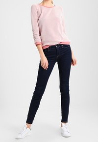 Suéter de punto de color rosa claro con mangas largas, combinado con jeans ajustados oscuros y zapatillas blancas con detalles verdes.