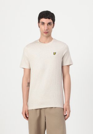 Giovane uomo con capelli corti e scuri che indossa una maglietta beige chiaro con un piccolo logo giallo a forma di uccello sul petto e pantaloni beige.