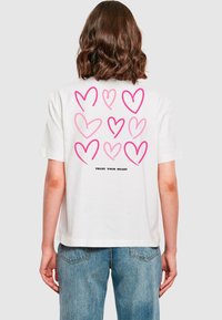 Weißes Baumwoll-T-Shirt mit einem rosa Herzmuster und dem Text "VERTRAUE DEINEM HERZEN" auf der Rückseite. Kurzärmlig, lockere Passform, schlichtes Design.