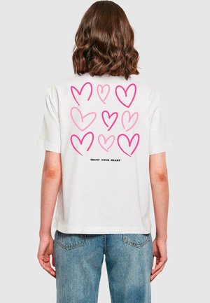 T-shirt bianco in cotone con un motivo a cuori rosa e la scritta "FIDATI DEL TUO CUORE" sul retro. Maniche corte, vestibilità comoda, design semplice.