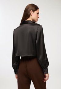 Przycięta czarna satynowa bluzka z długimi bufkami i klasycznym kołnierzykiem. Gładka tekstura, zapinane mankiety i prosta dolna krawędź.