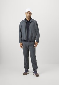 Mann iført en mørkegrå jakke og bukser, svart skjorte, hvit caps og lilla joggesko, stående mot en enkel lys bakgrunn.