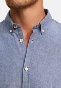 Lyseblå chambray-skjorte med en krave med knapper, beige knapper og en tekstureret vævning, med et delvist udsyn til halsudskæringen.