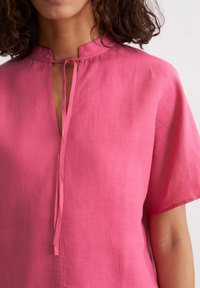 Kurzärmlige, pinke Leinenbluse mit einem Schlüsselloch-Ausschnitt und Bindendetail. Weiche Textur mit lockerem Schnitt und leichtem Fall.