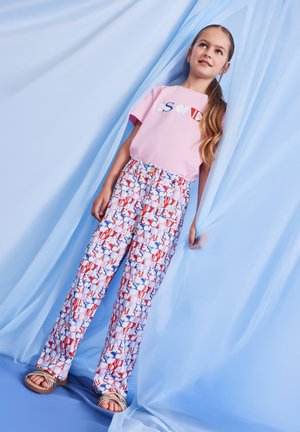 Ragazza giovane in piedi che tiene tende azzurro chiaro, indossa una maglietta rosa e pantaloni multicolore con motivo di testo ripetuto, sandali beige.