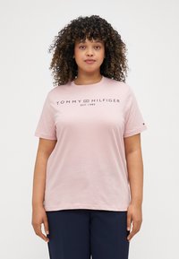 Svetloružový bavlnený tričko s krátkymi rukávmi a okrúhlym výstrihom. Na hrudi sa nachádza logo "TOMMY HILFIGER" v námorníckej modrej farbe.