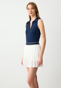 Marineblauwe mouwloze polo met V-hals en een witte geplooide rok, met een contrasterende tailleband met blauwe strepen. Glad textuur.
