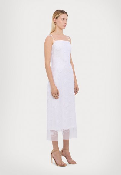 Vestido midi blanco bordado con tirantes finos, que presenta patrones florales, un dobladillo inferior transparente y se acompaña de sandalias de tacón alto beige.