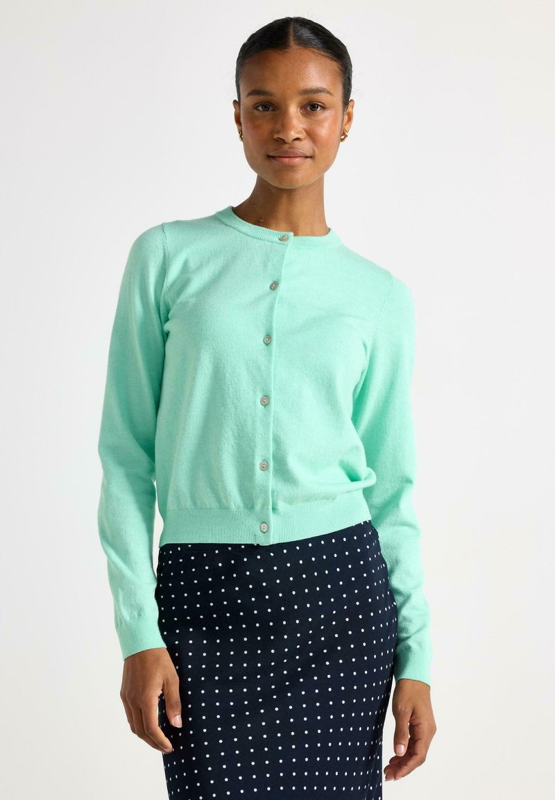 Mintgrön cardigan i mjukt material, med rund halsringning, knappstängning och ribbade ärmslut, parat med en prickig kjol.