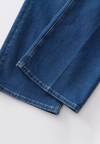 Jean en denim bleu moyen, avec une texture lisse et un design à jambes droites. Les ourlets sont soigneusement finis avec des détails de couture subtils.