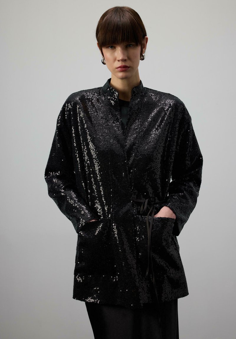 Žena s krátkymi hnedými vlasmi, oblečená v čiernom flitrovanom saku, s rukami v kapsách, stojaca pred jednoduchým sivým pozadím.