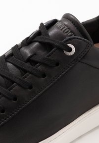 Sneaker in pelle nera con lacci piatti, dotata di occhielli metallici e una suola in gomma bianca. Texture liscia, design minimalista e dettaglio del logo.