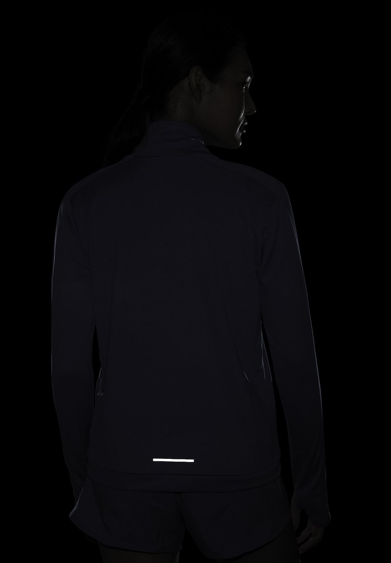 Chaqueta deportiva ajustada y oscura con mangas largas, cuello alto y detalles reflectantes en la parte inferior. Textura suave, diseñada para ser visible en condiciones de poca luz.