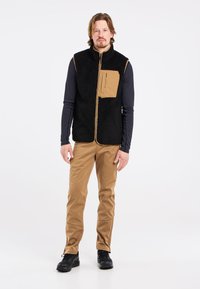 Juodas mikrofliso liemenė su smėlio spalvos priekinė kišenė ir užtrauktukas, apsirengta virš ilgos rankovės marškinėlių, derinama su smėlio spalvos kelnėmis ir juodais batais.