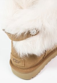 Bottine en daim couleur camel avec un revers en fourrure blanche duveteuse, ornée d'un bouton décoratif et d'un patch logo de la marque sur le talon.