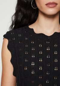 Černý háčkovaný top s vlnitým výstřihem a rukávy na kapesníky, s texturovaným vzorem otevřených vazeb a květinovými motivy.