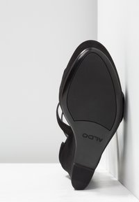 Chaussure plate noire à bout pointu, semelle texturée et nom de la marque ALDO sous la chaussure, appuyée contre un angle blanc entre un mur et le sol.