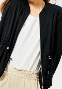 Svart bombardjacka med frontdragkedja, elastiska ärmar och dragsko i nederkant. Bärs över en vit skjorta, i kombination med ljusbeiga byxor.