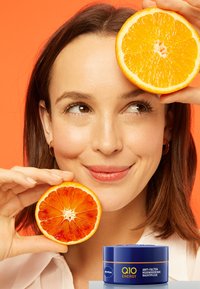 Blauer Nivea Q10 Energy Cream-Glas am Boden, Frau mit geschnittenen Orangen und Blutorangen auf einem lebhaften orangefarbenen Hintergrund. Glatte Haut, natürliches Make-up.