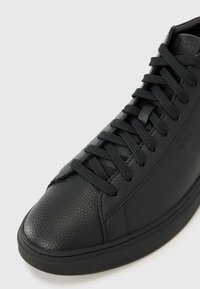 Zapatilla de piel negra con una superficie texturizada, punta redonda, cordones planos negros y suela de goma. Presenta una marca discreta.