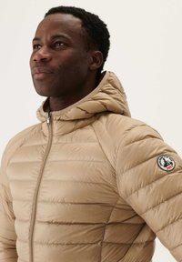 Beige puffjacka med huva, med quiltad struktur, framsida med dragkedja och en logotyp på vänster ärm. Lättviktig design.