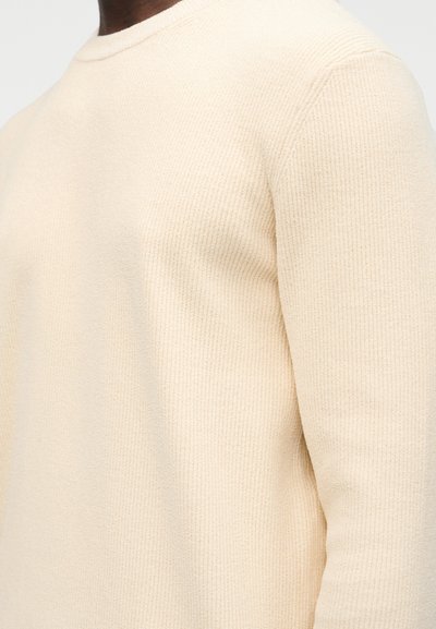 Gros plan sur un pull beige à manches longues côtelé porté par une personne, montrant la texture et les détails du tissu.