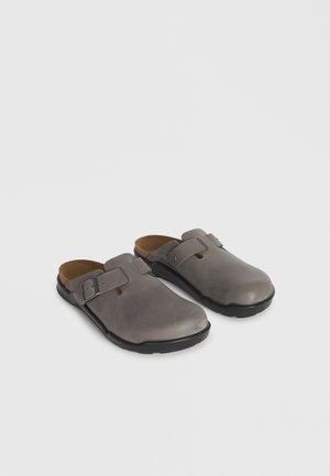 Par grå slip-on træsko med justerbare spænder og sorte såler, vist på en neutral lysegrå baggrund.