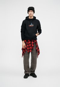 Crna dukserica s grafičkim printom, sivi cargo hlače i crveno karirano flanel košulja vezana oko struka. Crna kapa i crne tenisice.