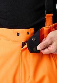 Oranje werkbroek met verstelbare zwarte bretels, ritssluiting en metalen drukknoopsluitingen. Glad materiaal dat functionaliteit en duurzaamheid benadrukt.
