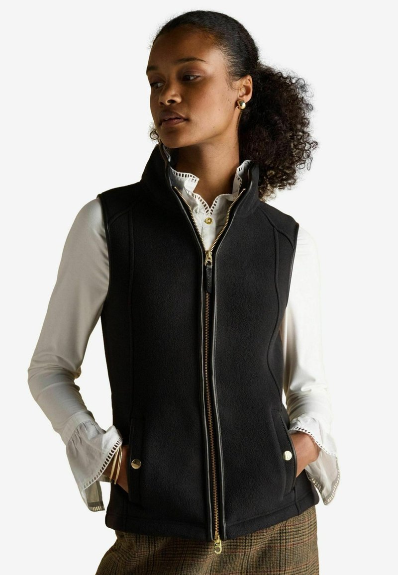 Čierna fleecová vesta s vysokým golierom, zlatým zipsom, bočnými vreckami a štruktúrovaným dizajnom, nosená cez bielu košeľu s volánovými manžetami.