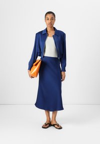 Giacca accorciata blu navy, canotta crema e gonna lunga blu. Tiene una borsa geometrica arancione. Infradito nere completano il look.