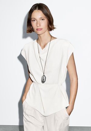 Weiße, drapierte Bluse mit Kappärmeln und einem V-förmigen Ausschnitt, kombiniert mit lockeren, hellen Hosen und einer großen silbernen Anhänger-Halskette.
