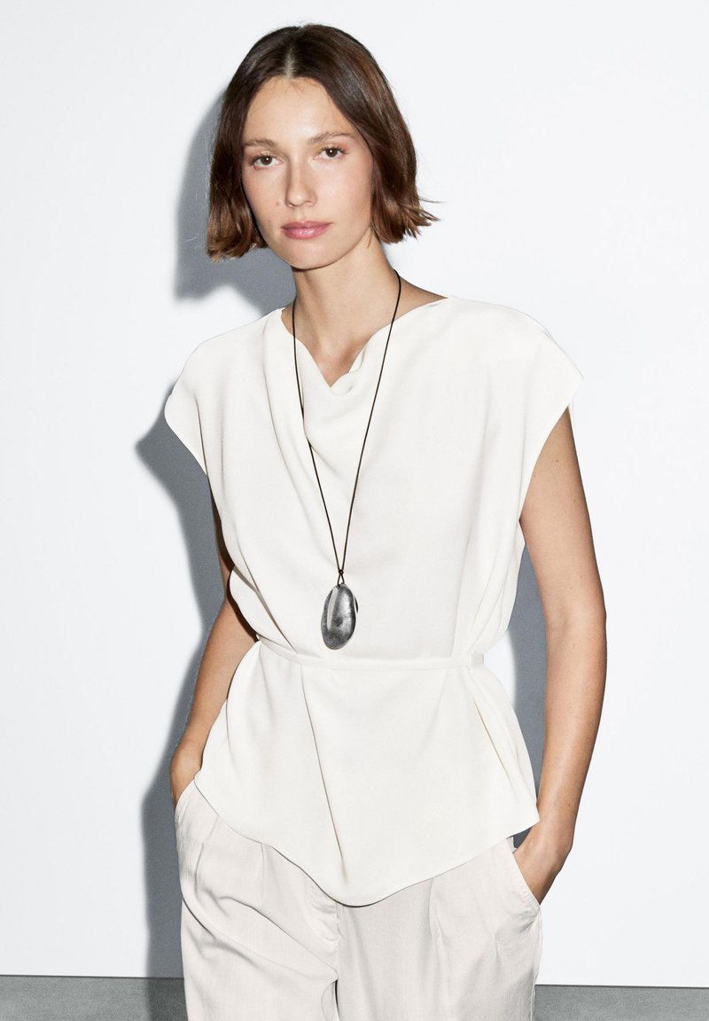 Witte, gedrapeerde blouse met kort kapmouwen en een V-hals, gecombineerd met losse, lichtkleurige broeken en een grote zilveren ketting met hanger.