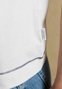 Marginea unei cămăși albe din bumbac, cu cusături bleu închis și o etichetă mică din material cu inscripția „Joules”, purtată cu blugi din denim albastru deschis.