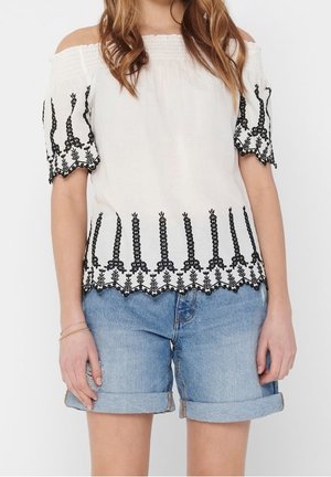 Witte off-shoulder blouse met zwarte bloemenborduursels, gedragen met opgerolde lichtblauwe denim shorts bij een staand persoon.