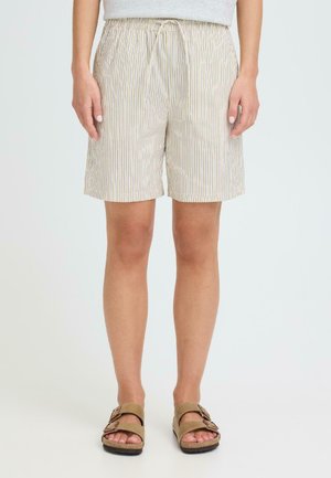 Osoba ubrana w beżowe, cienko prążkowane spodenki ze ściągaczem w pasie oraz sandały w kolorze brązowym z podwójną paską, stojąca na tle jednolitego, jasnego tła.