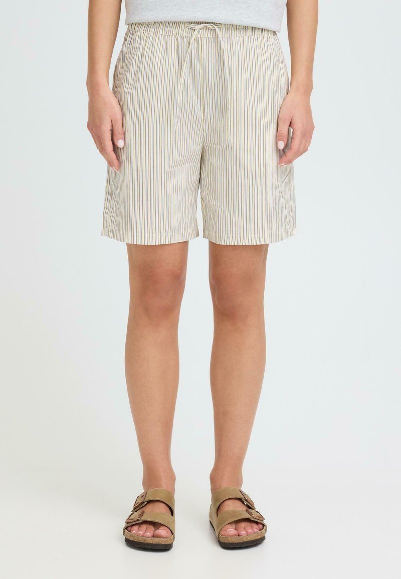 Άτομο που φοράει μπεζ, λεπτά ριγέ σορτς με κορδόνι στη μέση και ταμπά σανδάλια με διπλό λουρί, στέκεται μπροστά σε απλό ανοιχτό φόντο.